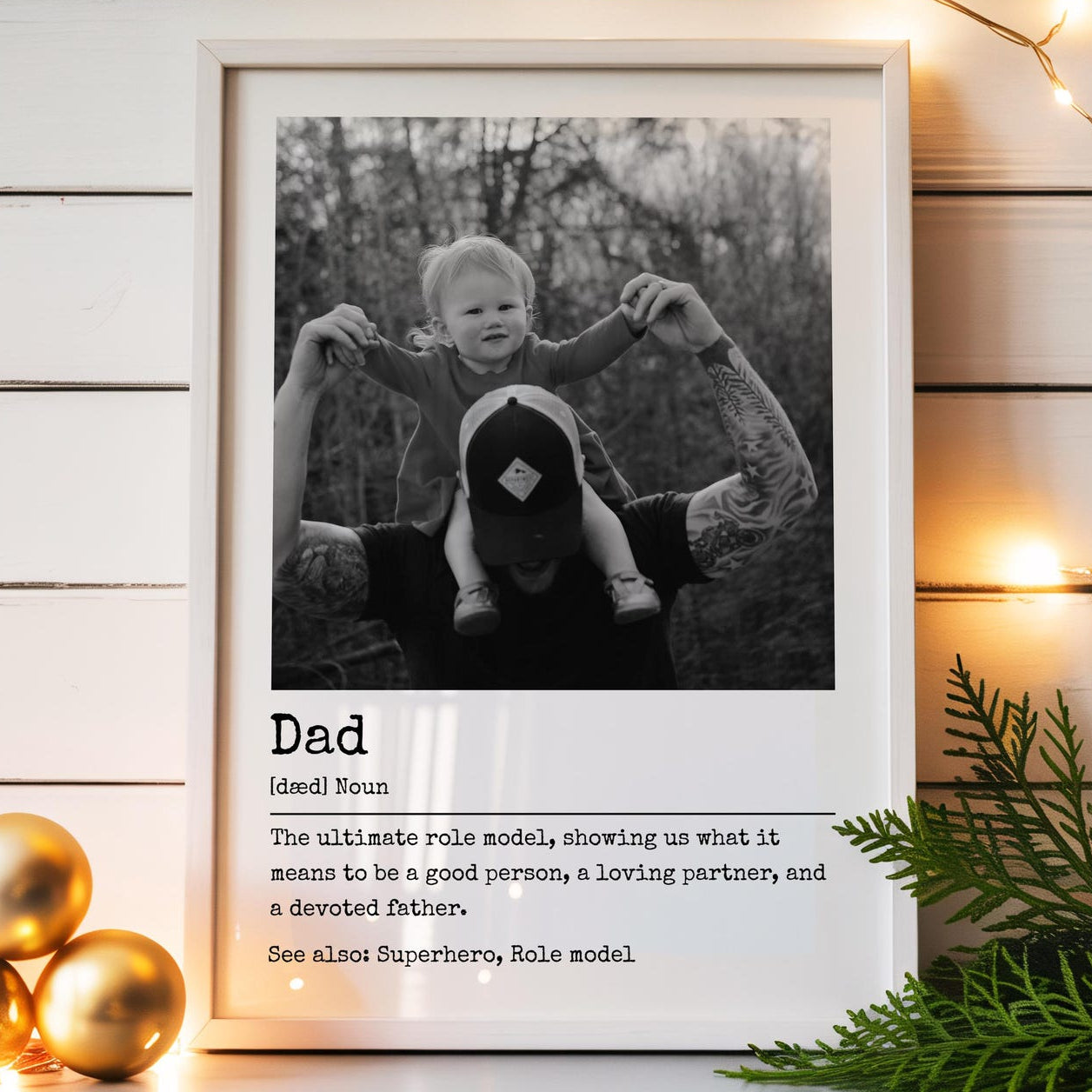 Framed photo of a child being held by an adult with a definition of 'Dad' below, on a wooden surface with decorative lights and ornaments.